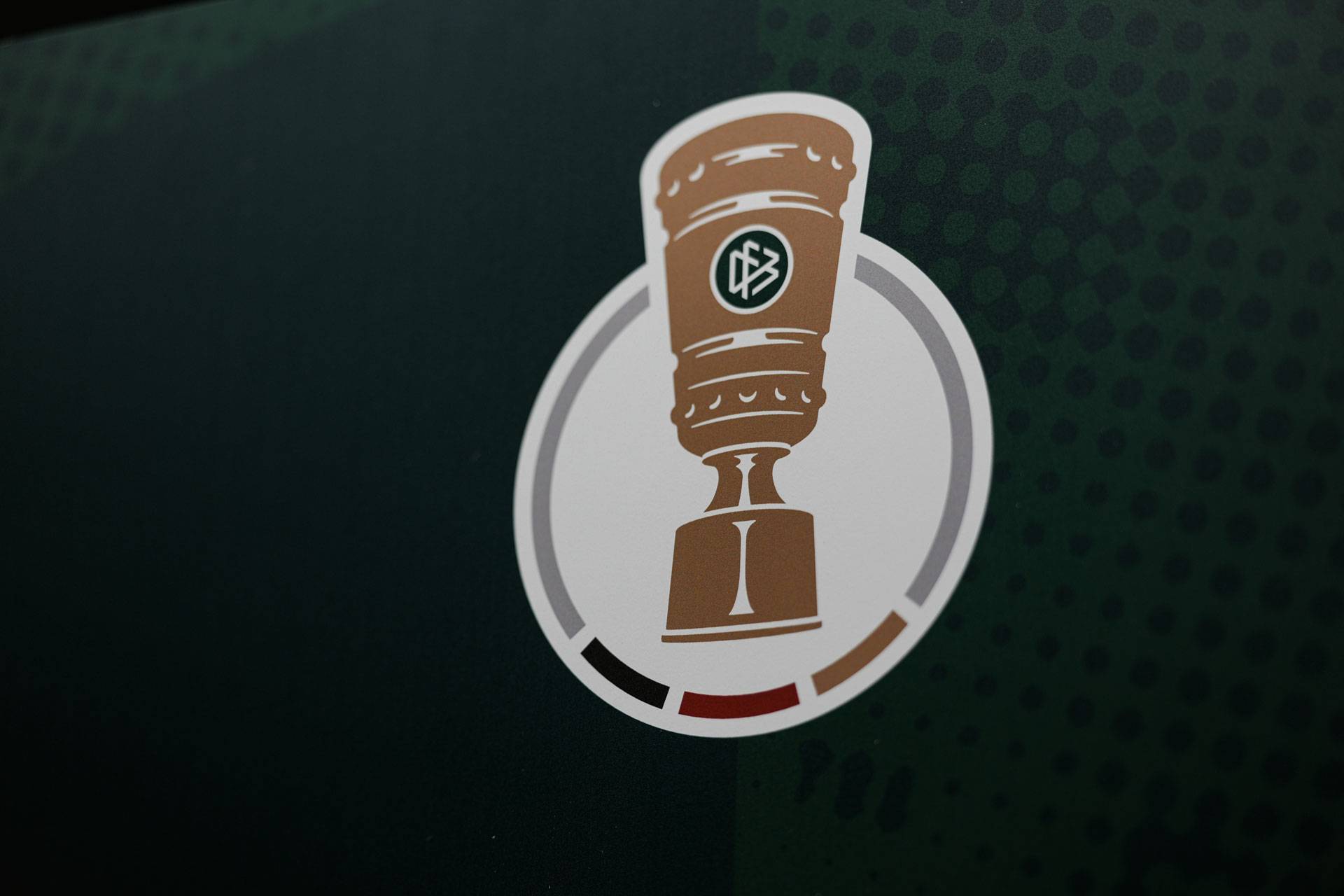 Logo einer braunen Tasse vor einem kreisförmigen Hintergrund mit grünen, roten und schwarzen Akzenten auf einem dunkelgrünen, gepunkteten Hintergrund.