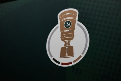 Logo eines Pokals vor einem kreisförmigen Hintergrund mit grünen, roten und schwarzen Akzenten auf einem dunkelgrünen, gepunkteten Hintergrund.