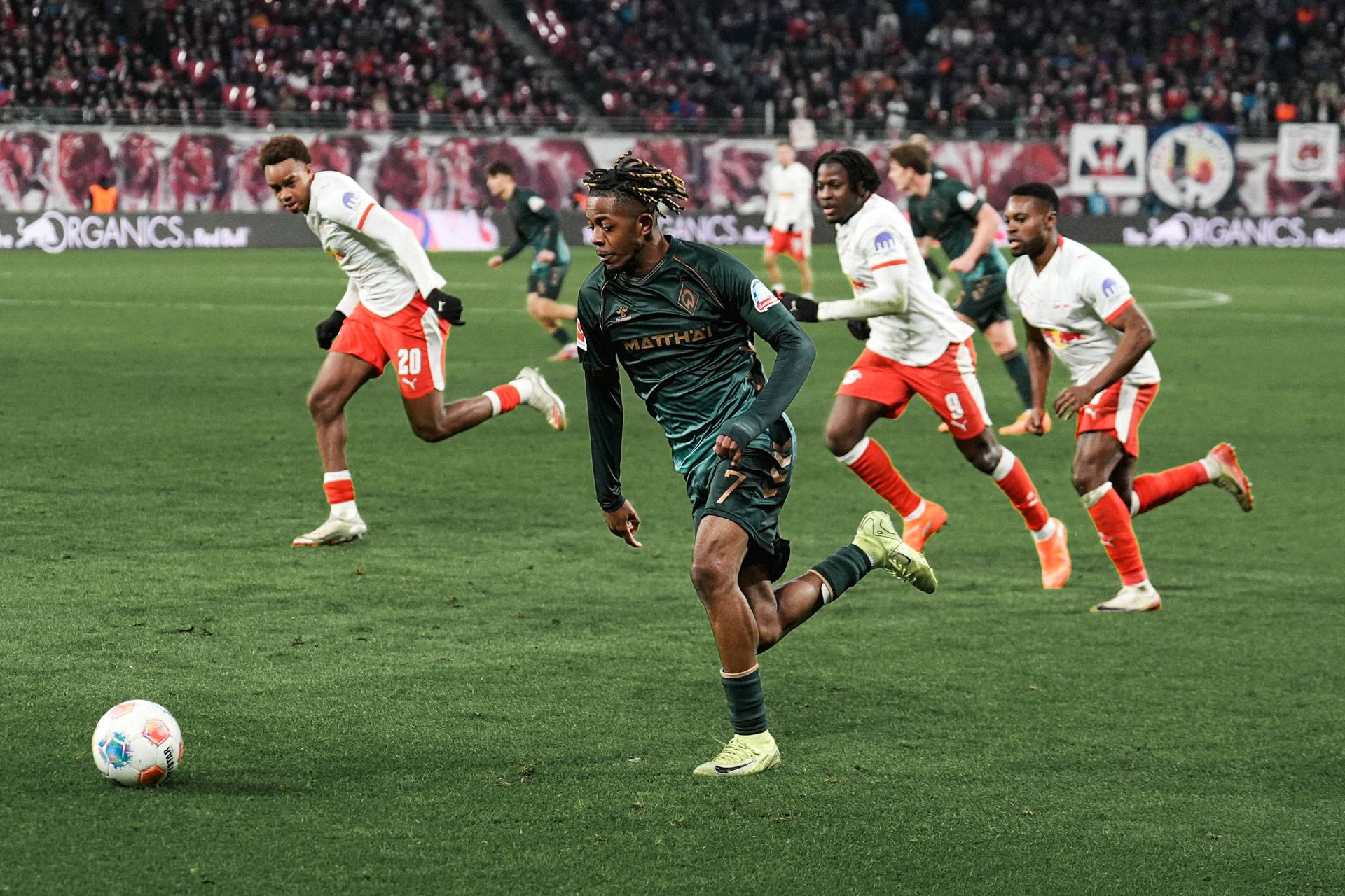 Ein Fußballspieler in grüner Kleidung dribbelt den Ball auf dem Spielfeld, verfolgt von Gegnern in weißen und roten Trikots, im Hintergrund ist eine Zuschauermenge zu sehen.