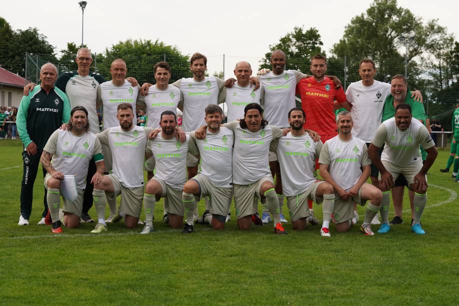 Eine Fußballmannschaft posiert auf einem Rasenfeld und trägt weiße Trikots mit grünen Akzenten. Im Hintergrund sind Bäume und Zuschauer zu sehen.