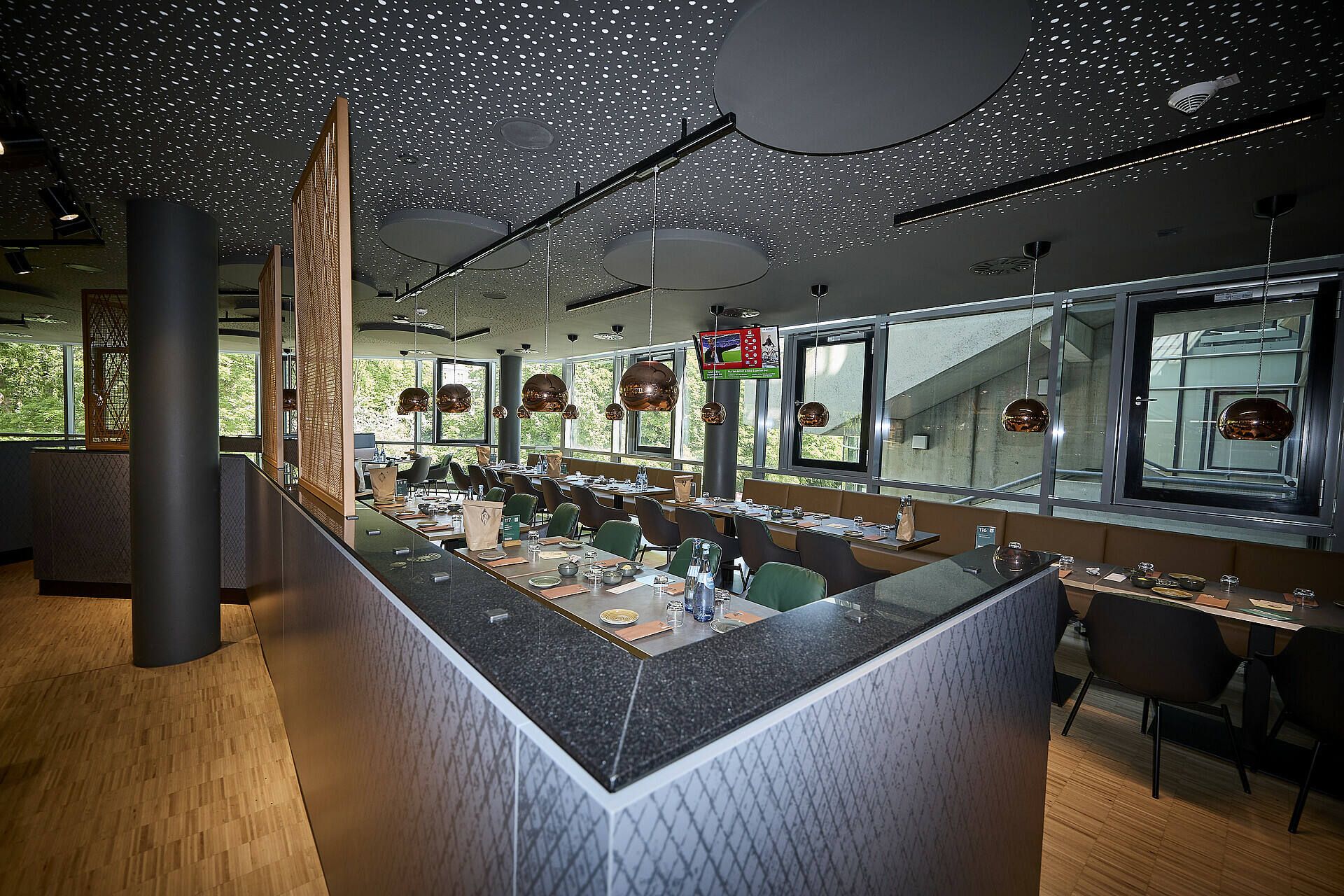 Modernes Restaurantinterieur mit Holzböden, dunklen Arbeitsplatten und kupferfarbenen Pendelleuchten, mit Tischen zum Essen in der Nähe großer Fenster.