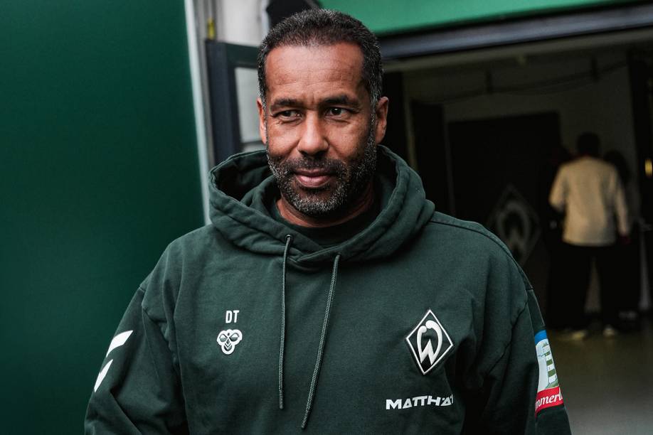 Ein Mann in einem grünen Sport-Kapuzenpulli mit Logos steht vor einem Gebäude.