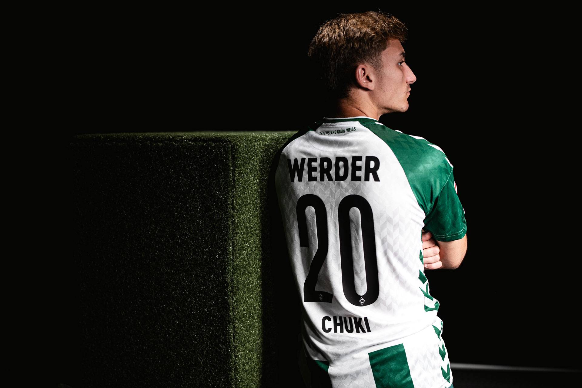 Fußballspieler in einem grün-weißen Werder-Trikot mit der Nummer 20 und dem Namen Chuki, der an einem grün strukturierten Block lehnt, im Seitenprofil.