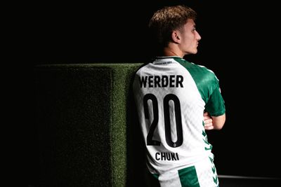 Fußballspieler in einem grün-weißen Werder-Trikot mit der Nummer 20 und dem Namen Chuki, der an einem grün strukturierten Block lehnt, im Seitenprofil.