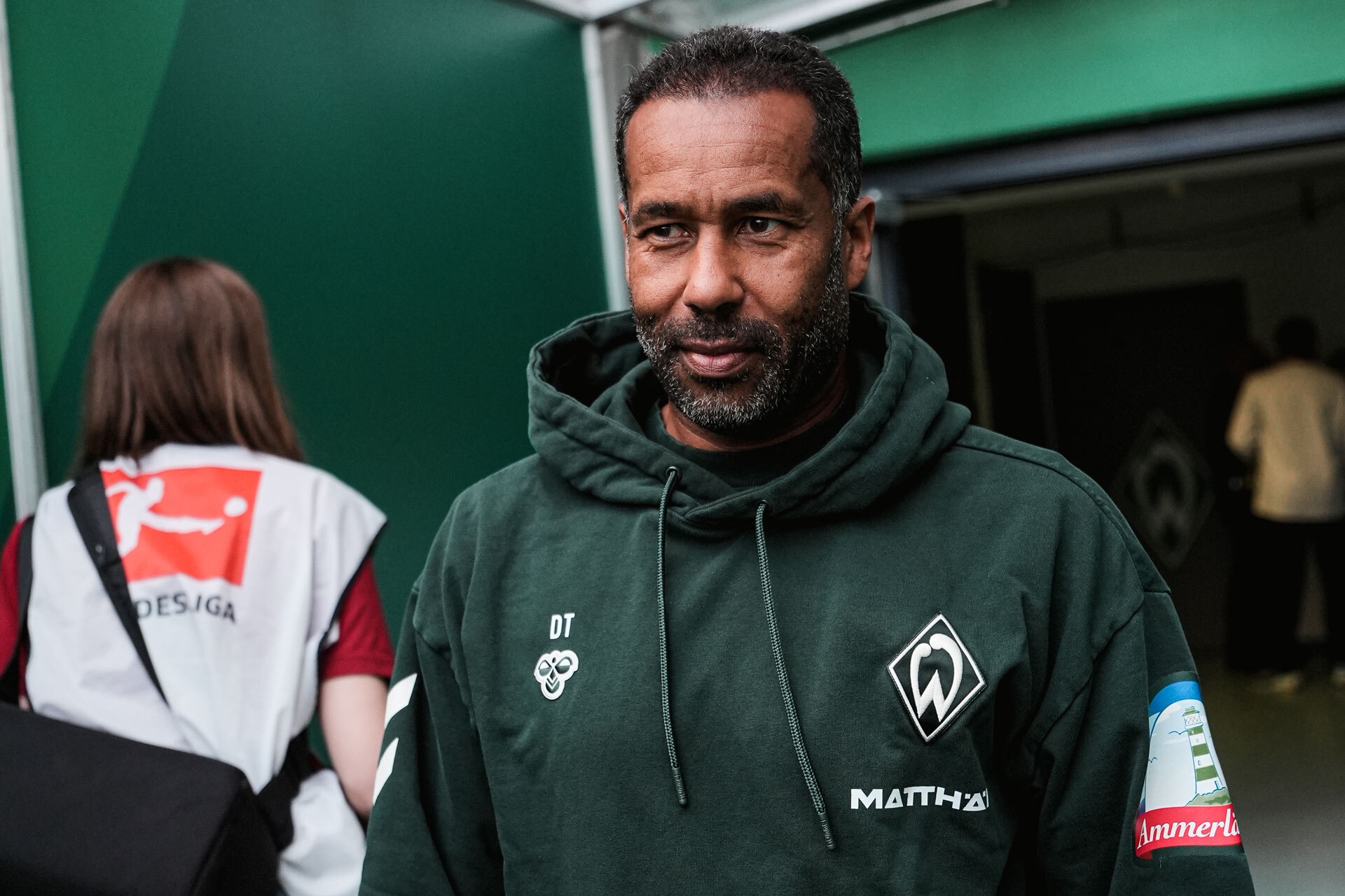 Ein Mann in einem grünen Kapuzenpullover mit Vereinslogos steht in einer Halle neben einer Frau mit langen Haaren, die eine Weste mit der Aufschrift „BUNDESLIGA“ trägt.
