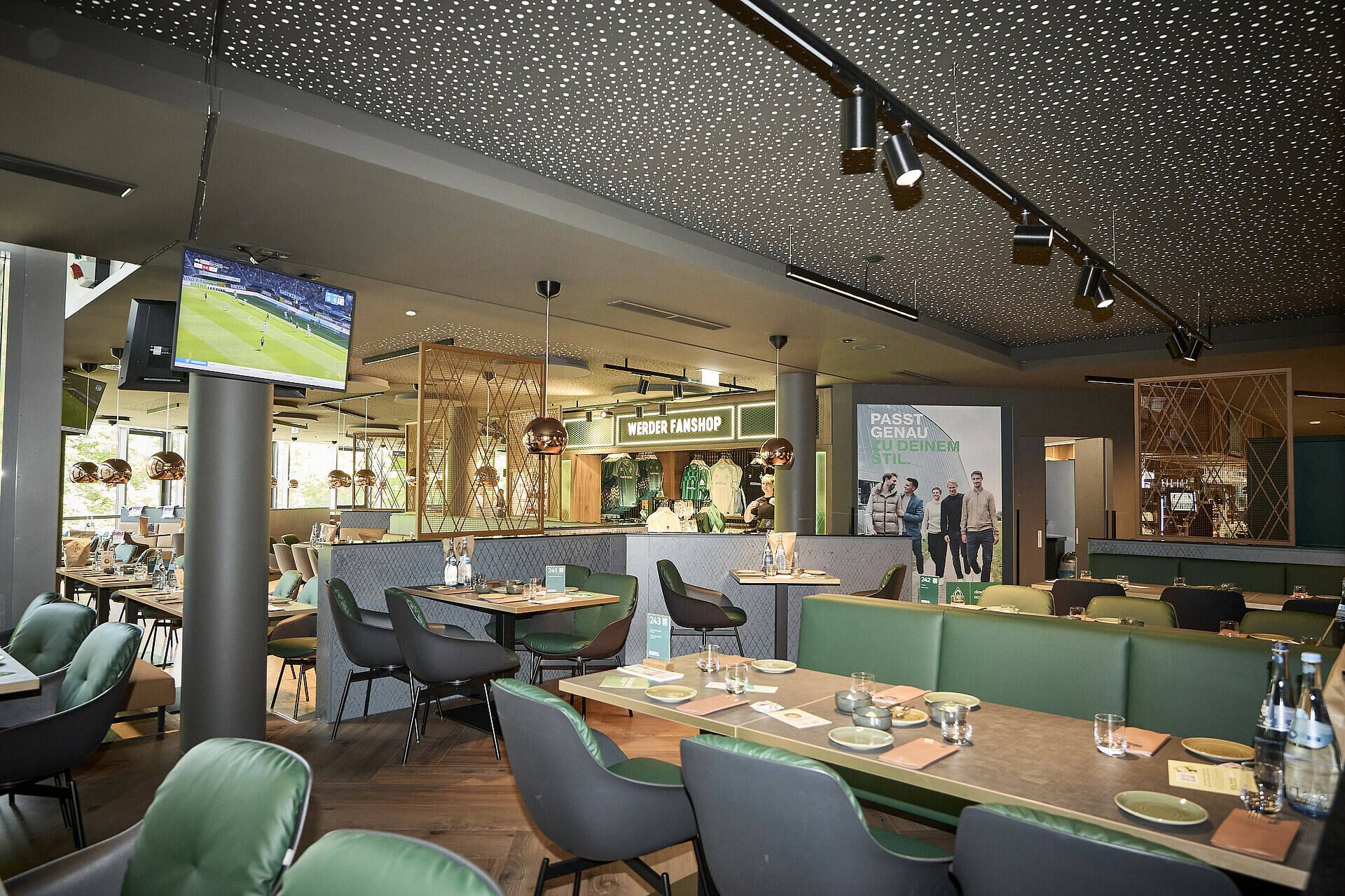 Modernes Restaurantinterieur mit grünen Sitzgelegenheiten, Holztischen mit Speisekarten und Besteck, einem Fernseher, auf dem ein Sportspiel läuft, und dekorativen Deckenleuchten.