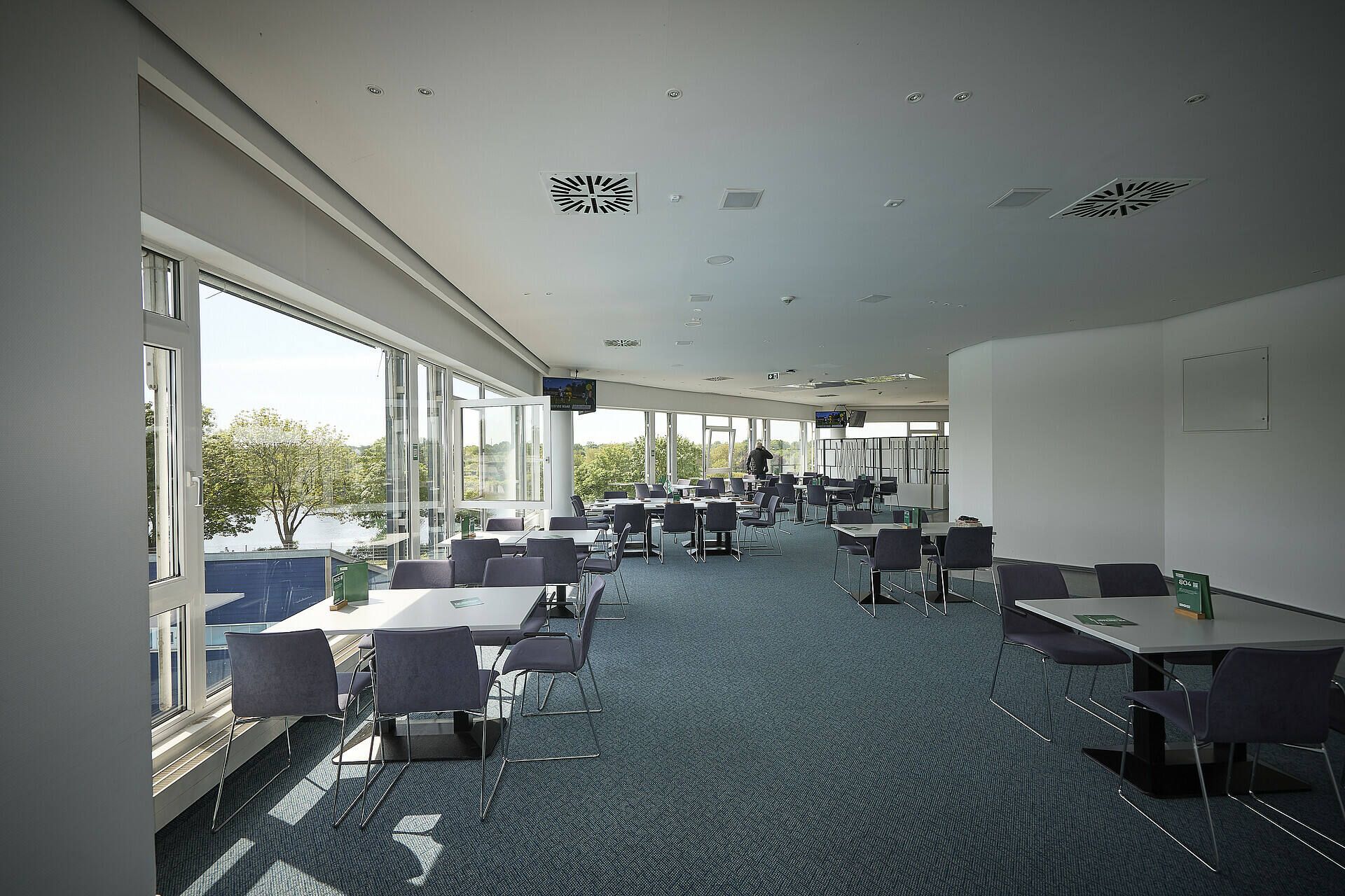 Geräumiger, moderner Speiseraum mit großen Fenstern, blauem Teppich und ordentlich angeordneten Tischen und Stühlen mit Blick auf eine malerische Außenlandschaft.