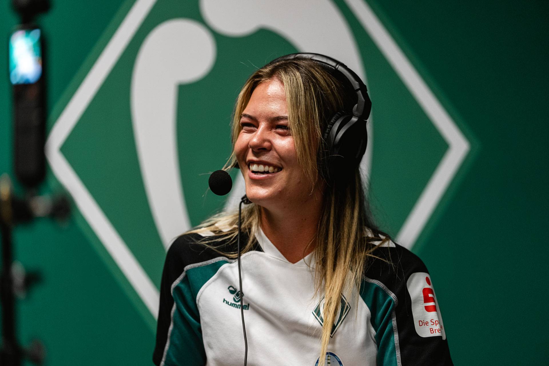 Eine lächelnde Frau mit Sportkopfhörern und einem grün-weißen Trikot spricht in ein Mikrofon, hinter ihr ist ein grünes Hintergrundlogo zu sehen.