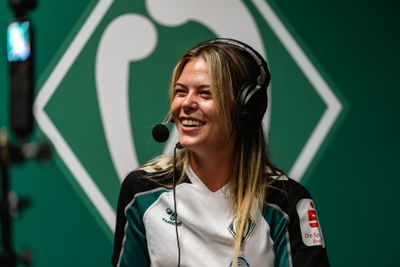 Eine lächelnde Frau mit Sportkopfhörern und einem grün-weißen Trikot spricht in ein Mikrofon, hinter ihr ist ein grünes Hintergrundlogo zu sehen.