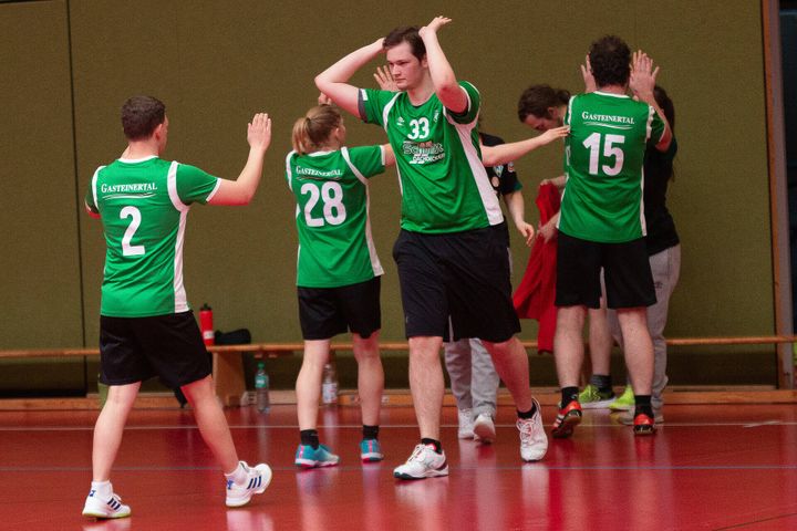 Eine Gruppe von Handballspielern in grünen Trikots klatschen sich auf einem roten Spielfeld ab und feiern oder fördern damit den Teamgeist.