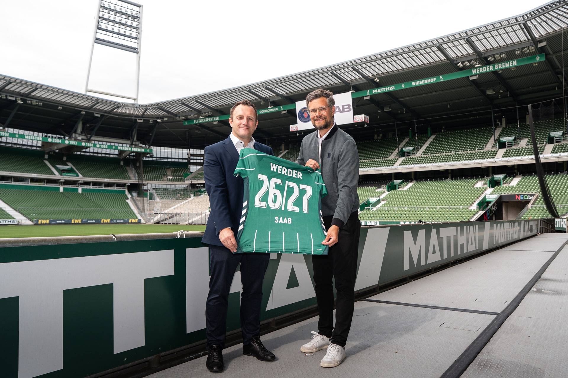 Zwei Männer, die ein grünes Werder-Bremen-Trikot mit der Aufschrift „26/27“ in den Händen halten und in einem leeren Fußballstadion stehen.