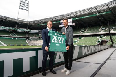 Zwei Männer, die ein grünes Werder-Bremen-Trikot mit der Aufschrift „26/27“ in den Händen halten und in einem leeren Fußballstadion stehen.