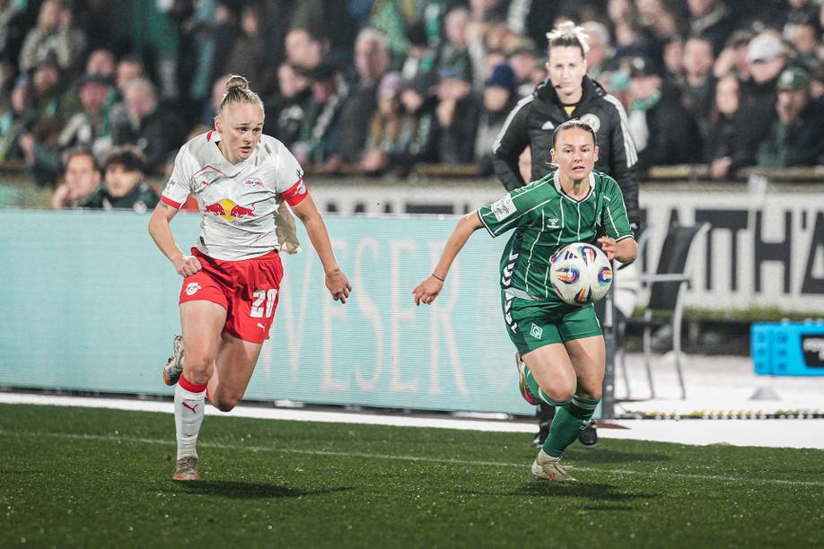 Zwei Fußballspielerinnen kämpfen auf dem Spielfeld um den Ball. Die eine trägt ein weiß-rotes Trikot, die andere ein grünes. Im Hintergrund sind ein Trainer und Zuschauer zu sehen.
