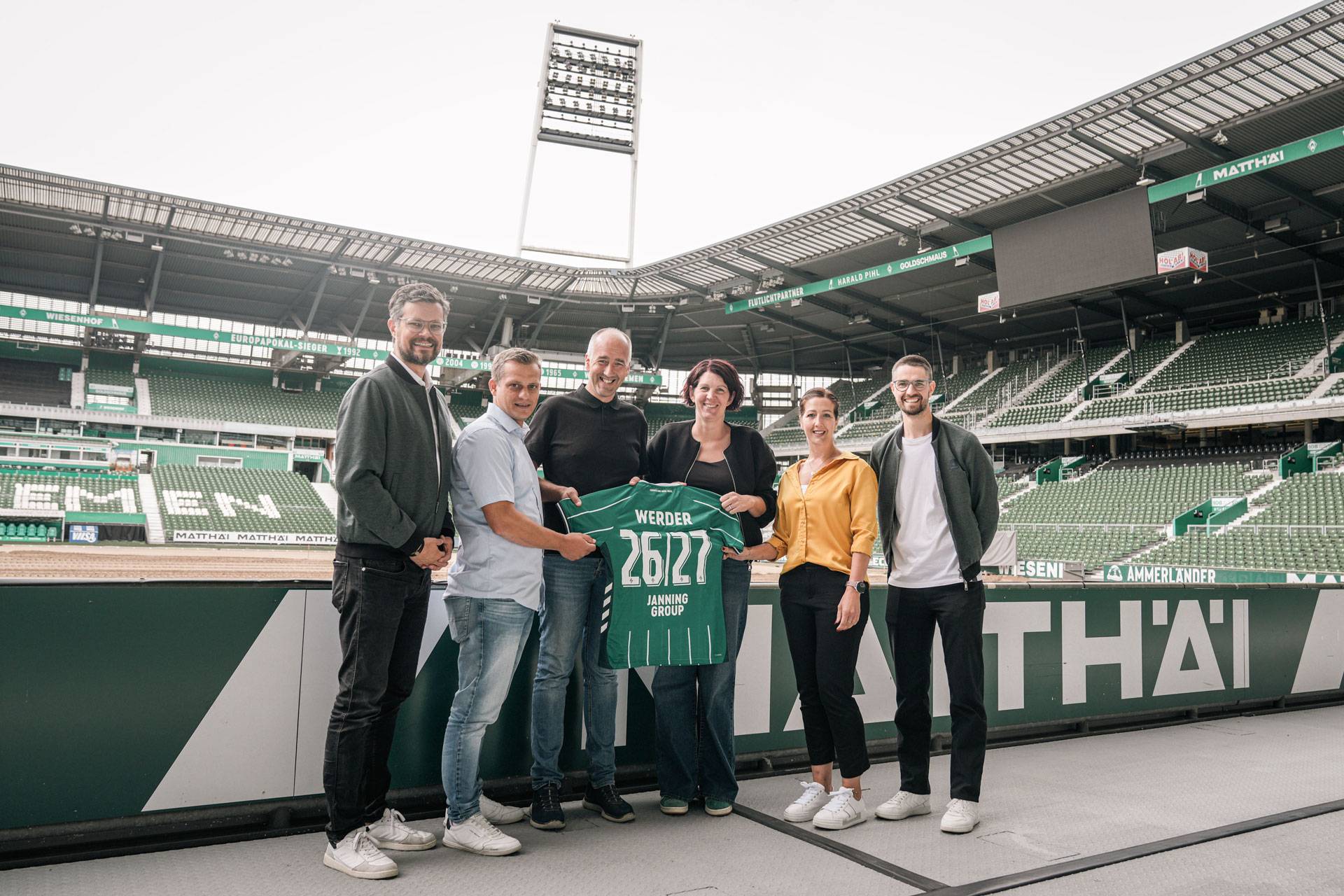 Sechs Personen stehen in einem Stadion und halten ein grünes Werder-Trikot mit der Nummer 26127 in den Händen.