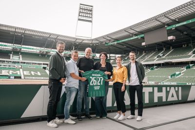 Sechs Personen stehen in einem Stadion und halten ein grünes Werder-Trikot mit der Nummer 26127 in den Händen.