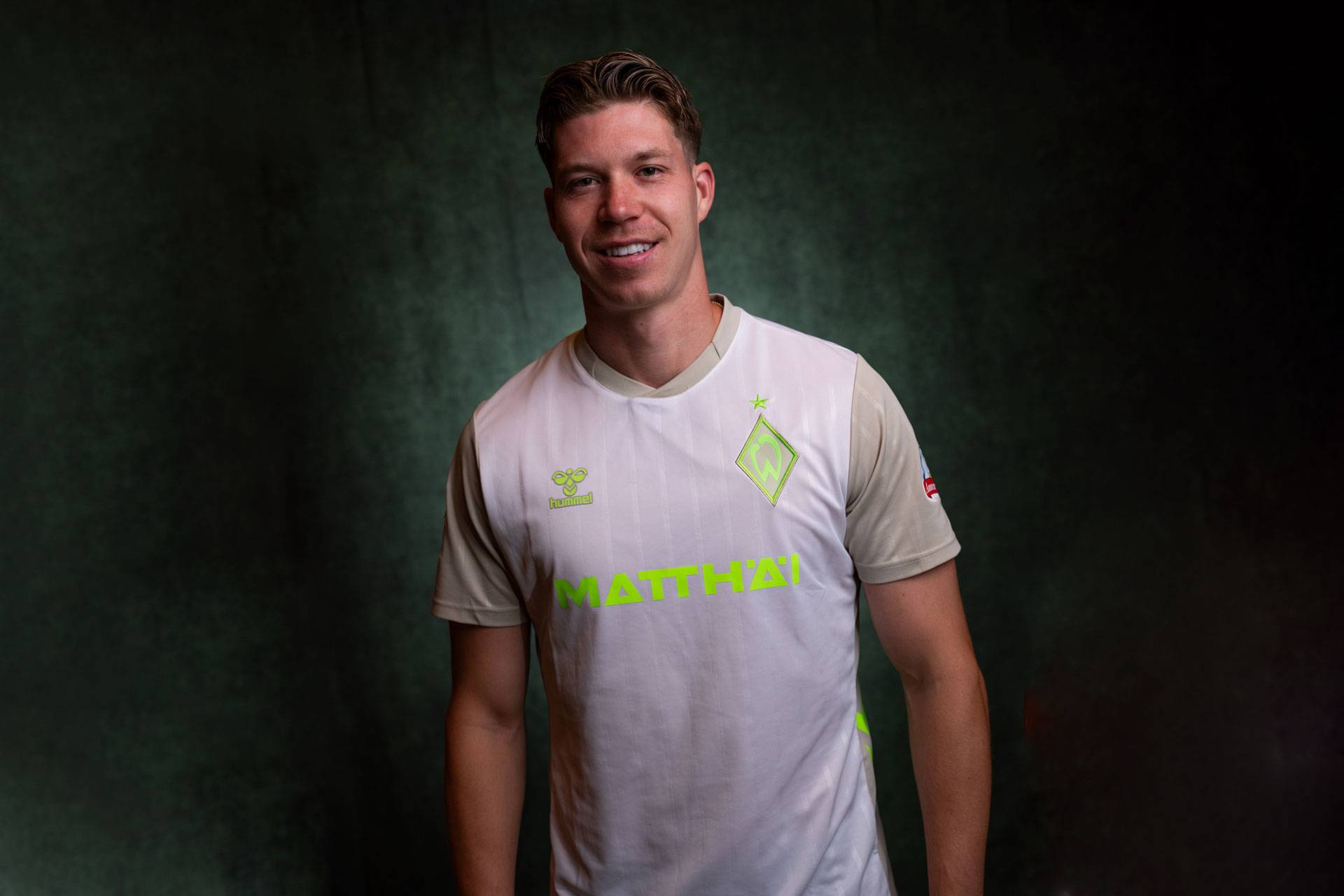 Lächelnder Mann in einem weiß-beigen Werder-Trikot vor einem dunklen Hintergrund.