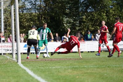 Szene aus einem Fußballspiel: Ein Spieler in Rot fällt in der Nähe des Tors, ein Torwart in grün-schwarzem Trikot und ein weiterer Spieler in grün-weiß gestreiftem Trikot.