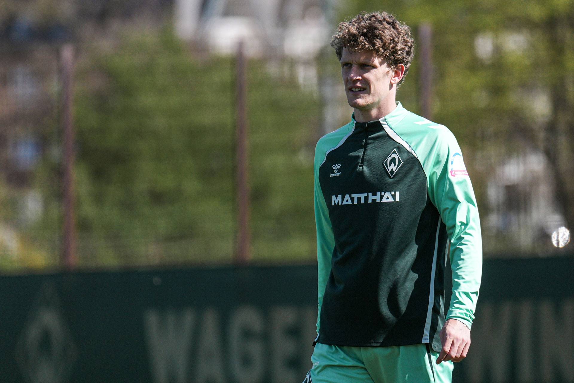 Ein Fußballspieler in grün-schwarzem Trainingsoutfit steht an einem sonnigen Tag im Freien, im Hintergrund sind verschwommene Bäume zu sehen.
