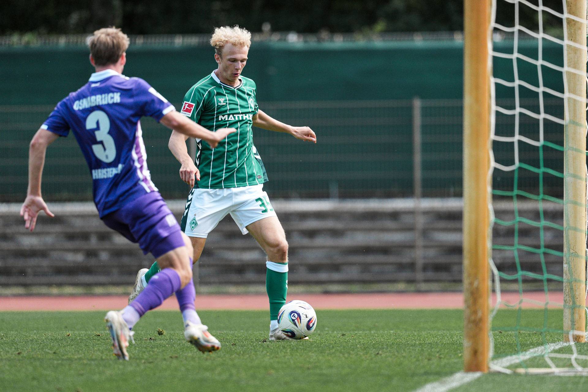 Ein Fußballspieler im grünen Trikot dribbelt in der Nähe des Tors, während ein anderer im violetten Trikot auf dem Rasen in die Verteidigung eilt.