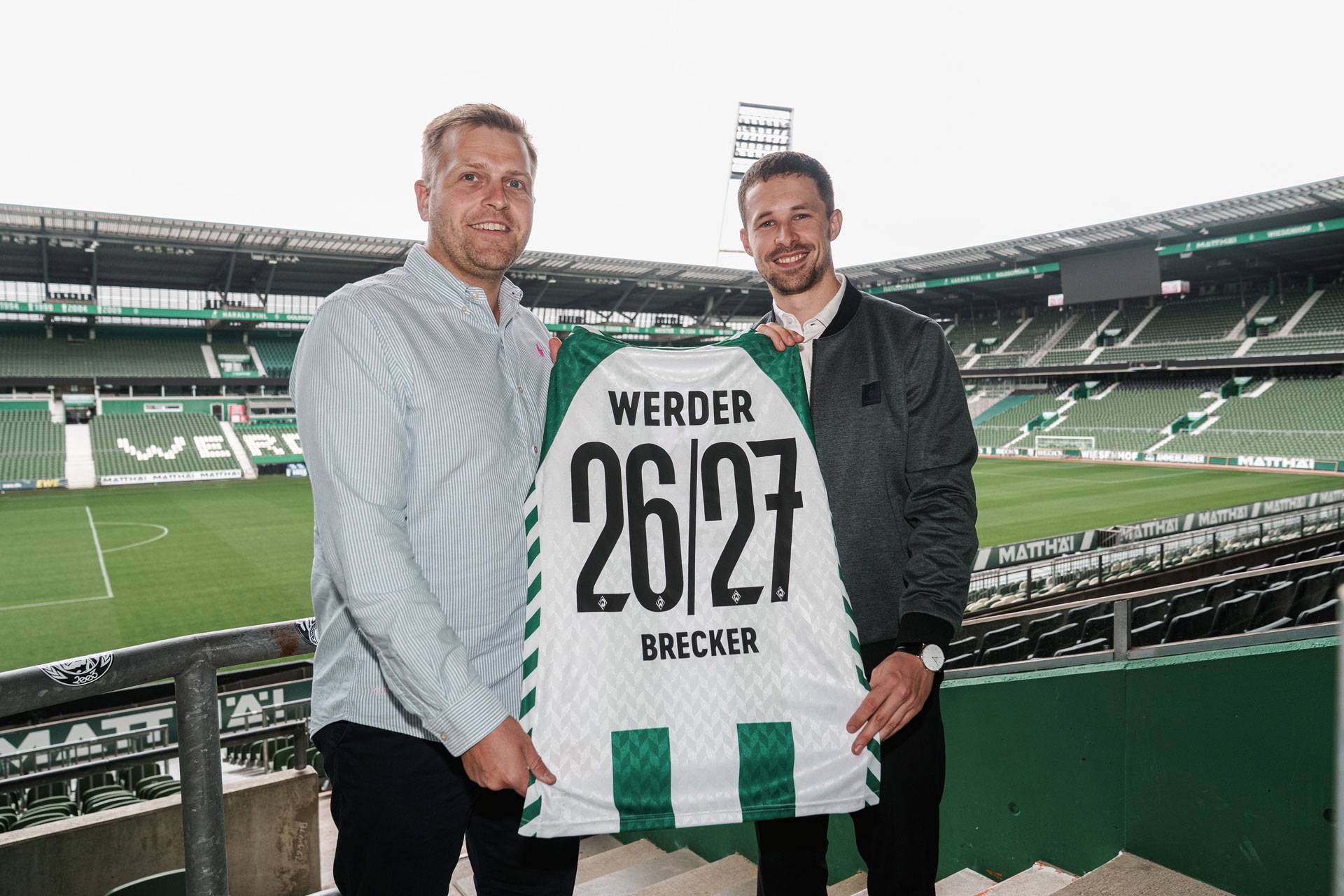 Zwei Personen, die ein Trikot von Werder Bremen mit der Aufschrift „26/27 Brecker“ in den Händen halten und in einem leeren Stadion stehen.