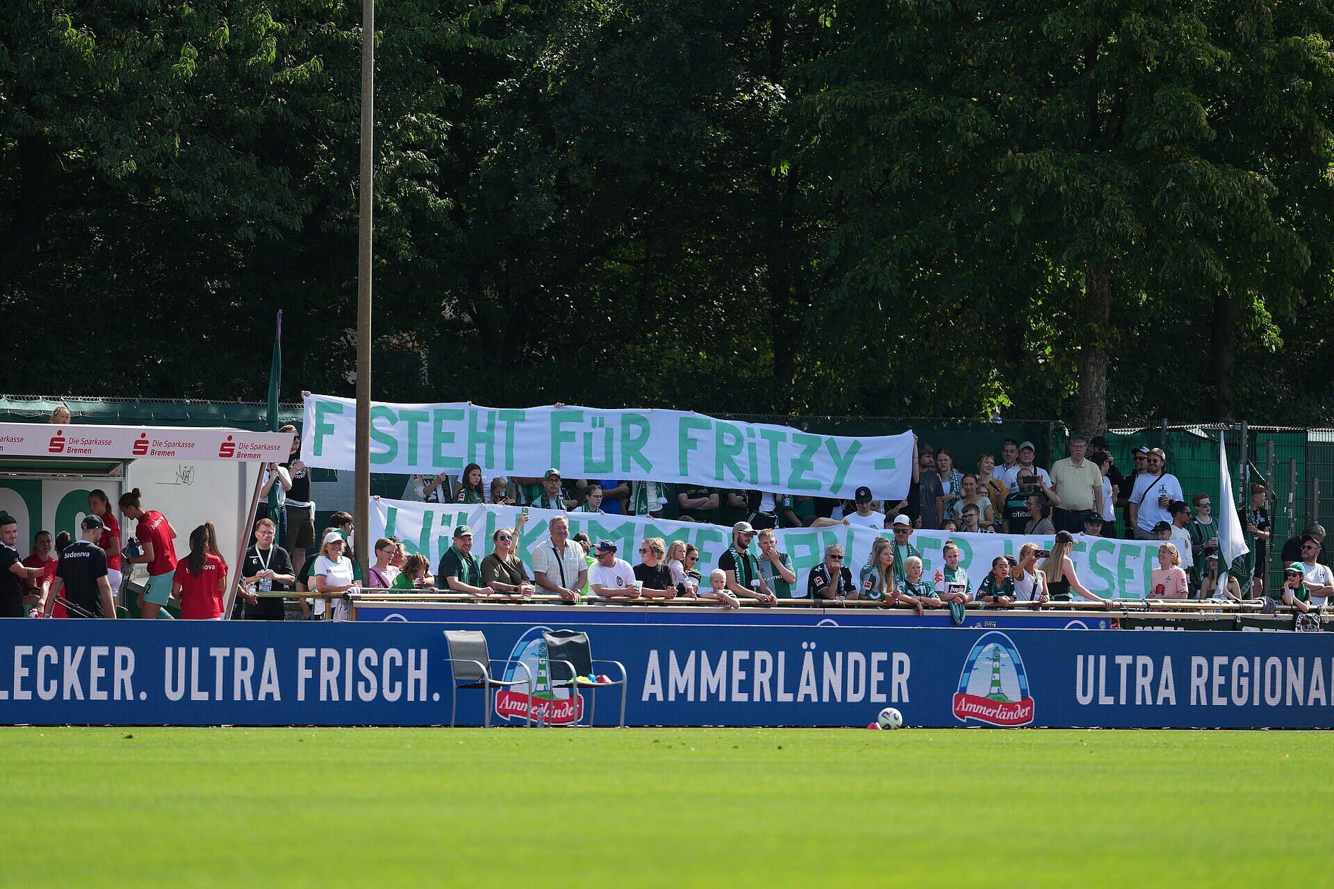 Fans halten ein Banner hoch mit dem Aufdruck: "F steht für Fritzy - Willkommen an der Weser". Im Hintergrund sind Bäume zu sehen. Die Fans stehen hinter einer Ammerländer-Werbebande am Spielfeldrand.
