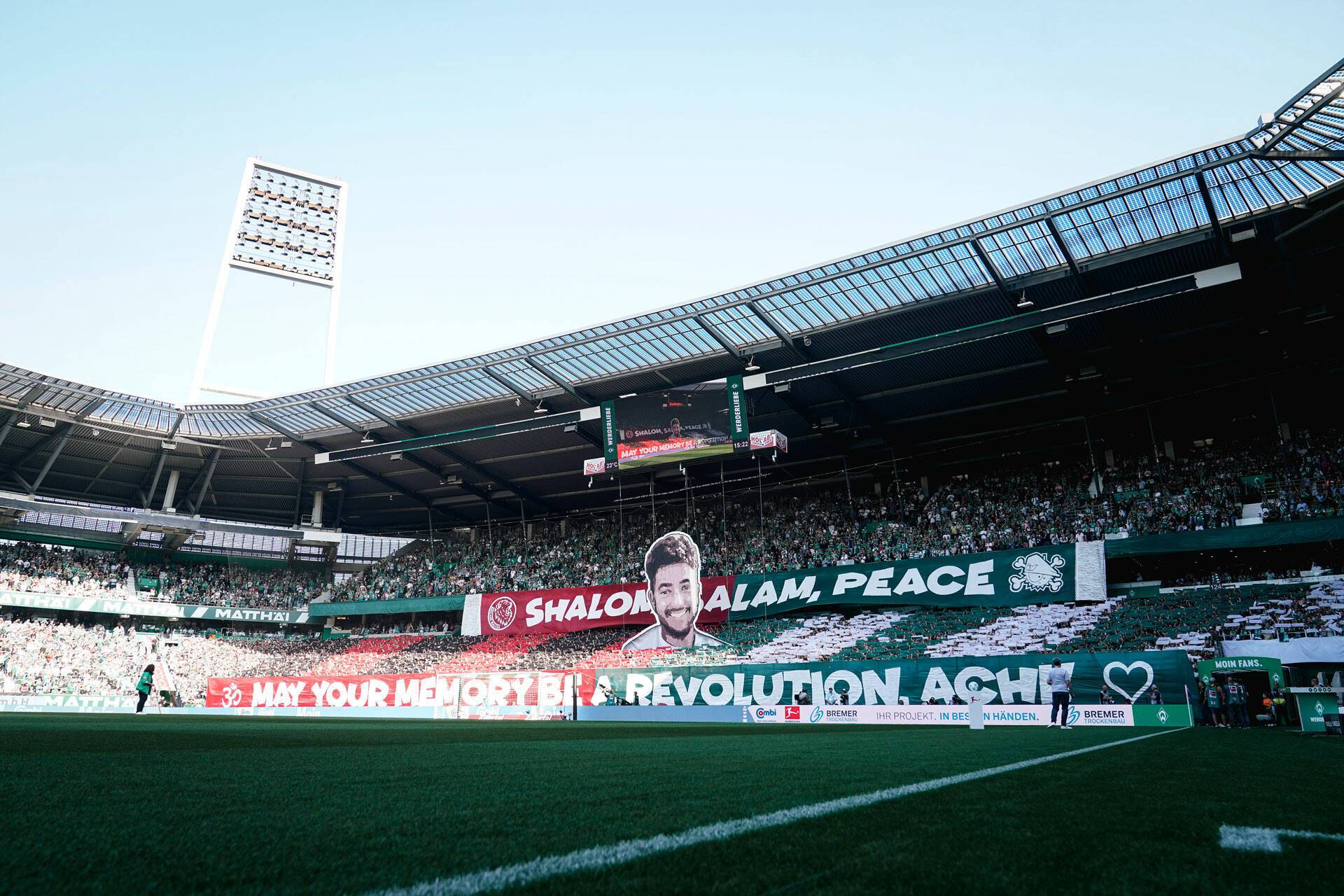 Ein Stadion voller Fans, die ein großes Banner mit dem Gesicht eines Mannes und den Worten „Shalom, Salam, Peace“ und „May Your Memory Be a Revolution, Ach!“ hochhalten.