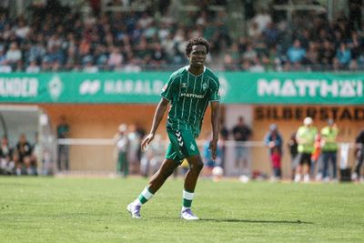 Fußballspieler im grünen Trikot auf dem Spielfeld während eines Spiels. Im Hintergrund sind Zuschauer auf den Tribünen und Regen zu sehen.