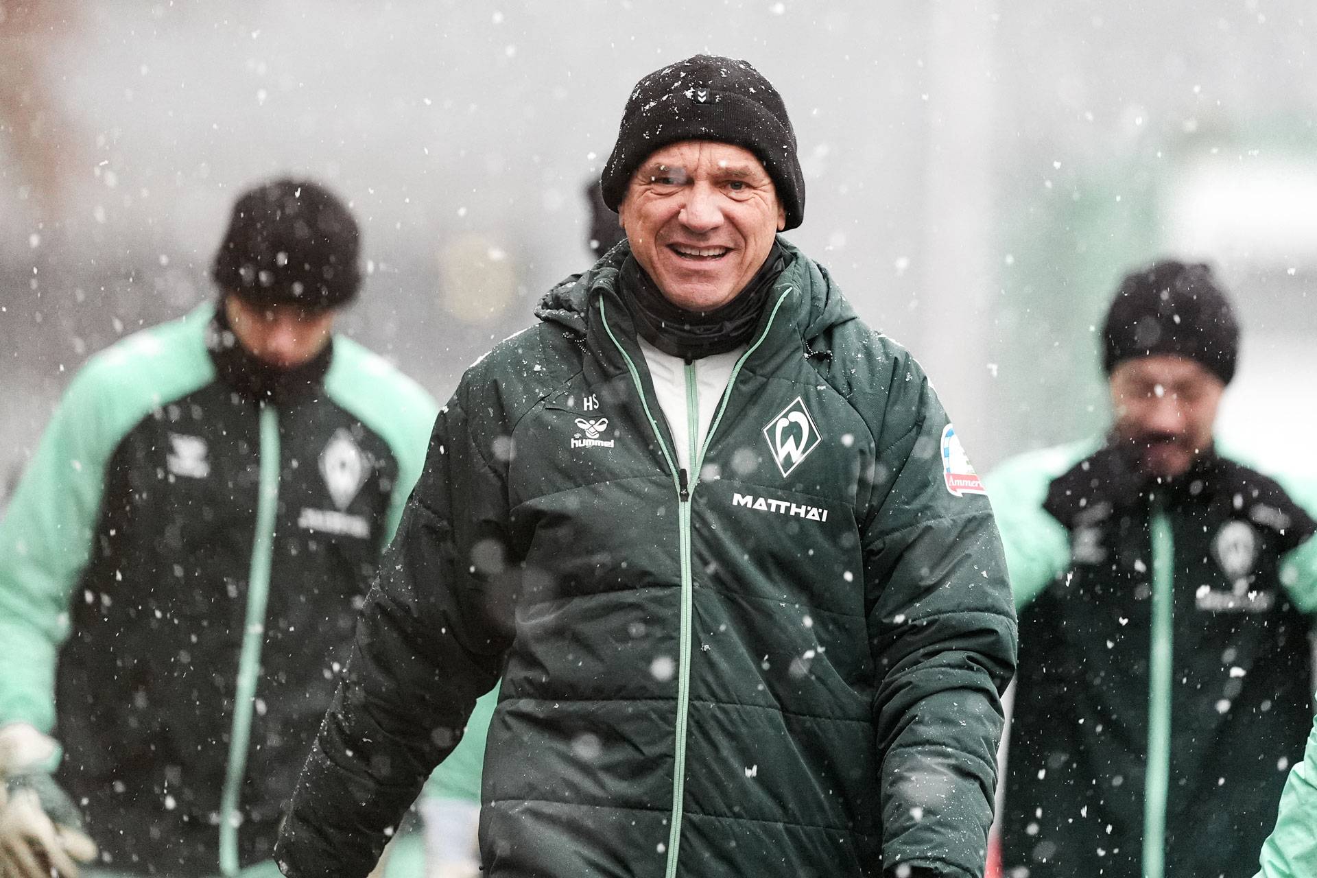 Horst Steffen in einer grünen Jacke und einer schwarzen Mütze lächelt bei Schneefall, im Hintergrund stehen zwei weitere Personen in ähnlicher Kleidung.