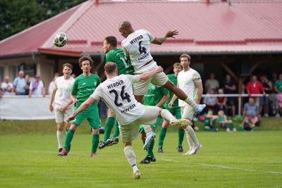 Fußballspieler in grün-weißen Trikots springen in der Nähe des Tores nach dem Ball, während im Hintergrund Zuschauer zuschauen.