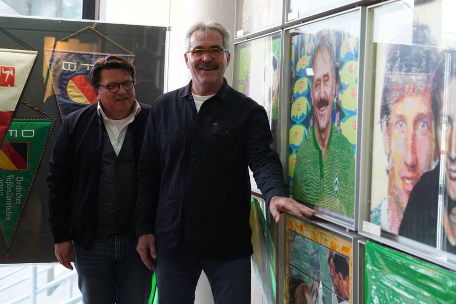 Zwei Männer stehen lächelnd vor einer Wand, an der in einer Ausstellungsumgebung farbenfrohe Porträts und Sport-Erinnerungsstücke zu sehen sind.