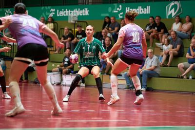 Hallenhandballspiel mit einem Spieler in einem grünen Trikot, der den Ball hält und seinen Gegnern in violetten Trikots gegenübersteht; Zuschauer beobachten das Spiel von der Tribüne aus.