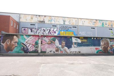 Ein farbenfrohes Wandbild an einer Hauswand zeigt Fußballspieler in dynamischen Posen, umgeben von Graffiti und urbaner Kunst.
