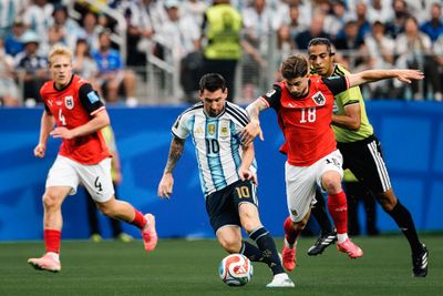 Fußballspieler aus Argentinien und Österreich kämpfen während eines Spiels um den Ball, im Hintergrund ist ein Schiedsrichter in einem gelben Trikot zu sehen.