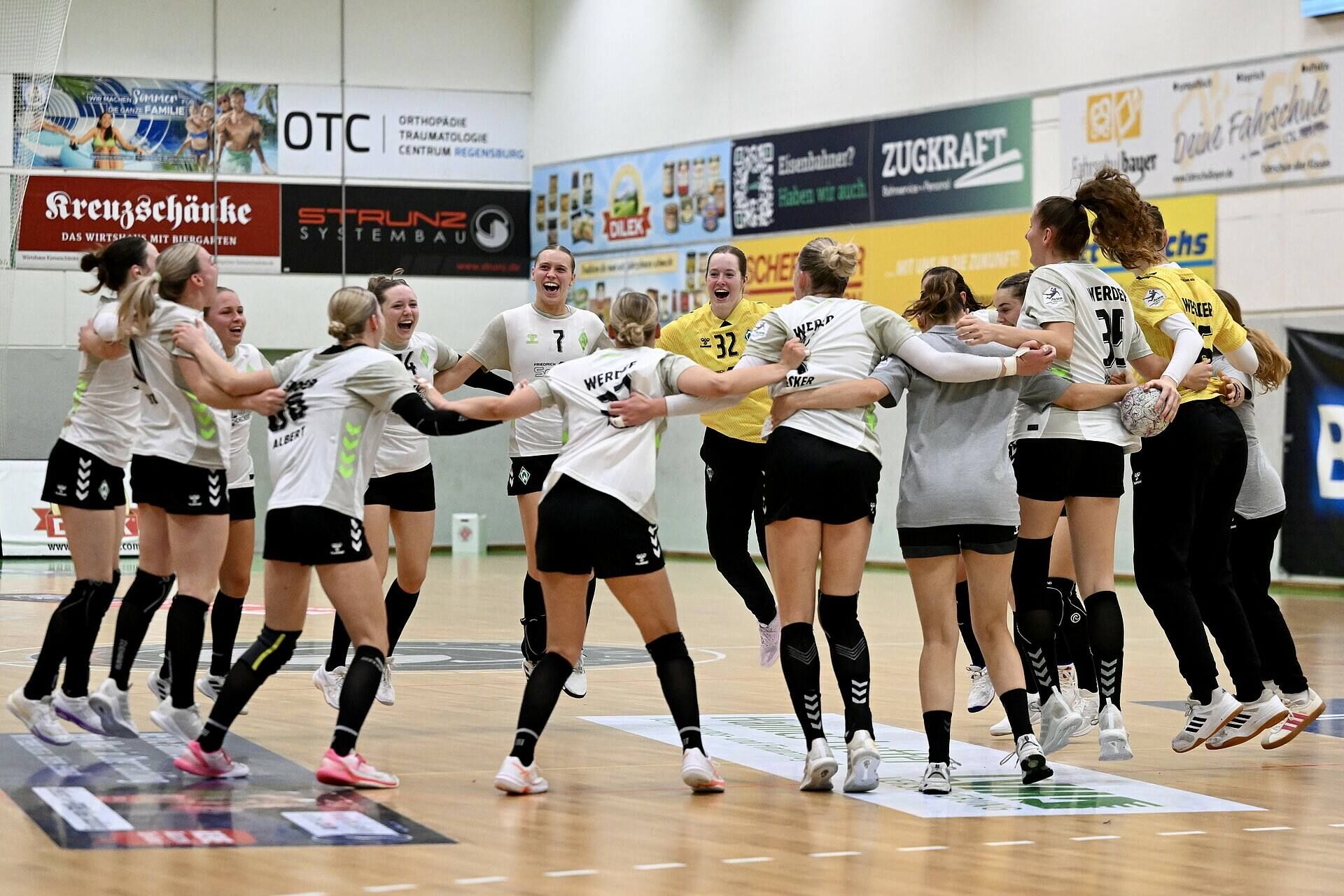 Eine Frauenhandballmannschaft feiert ausgelassen in einer Sporthalle, bildet einen Kreis und springt gemeinsam in die Luft. Im Hintergrund sind Werbebanner zu sehen.