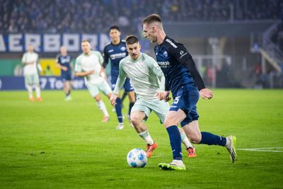 Zwei Fußballspieler kämpfen während eines Spiels auf einem grünen Rasen um den Ball, im Hintergrund sind mehrere Spieler und Zuschauer zu sehen.