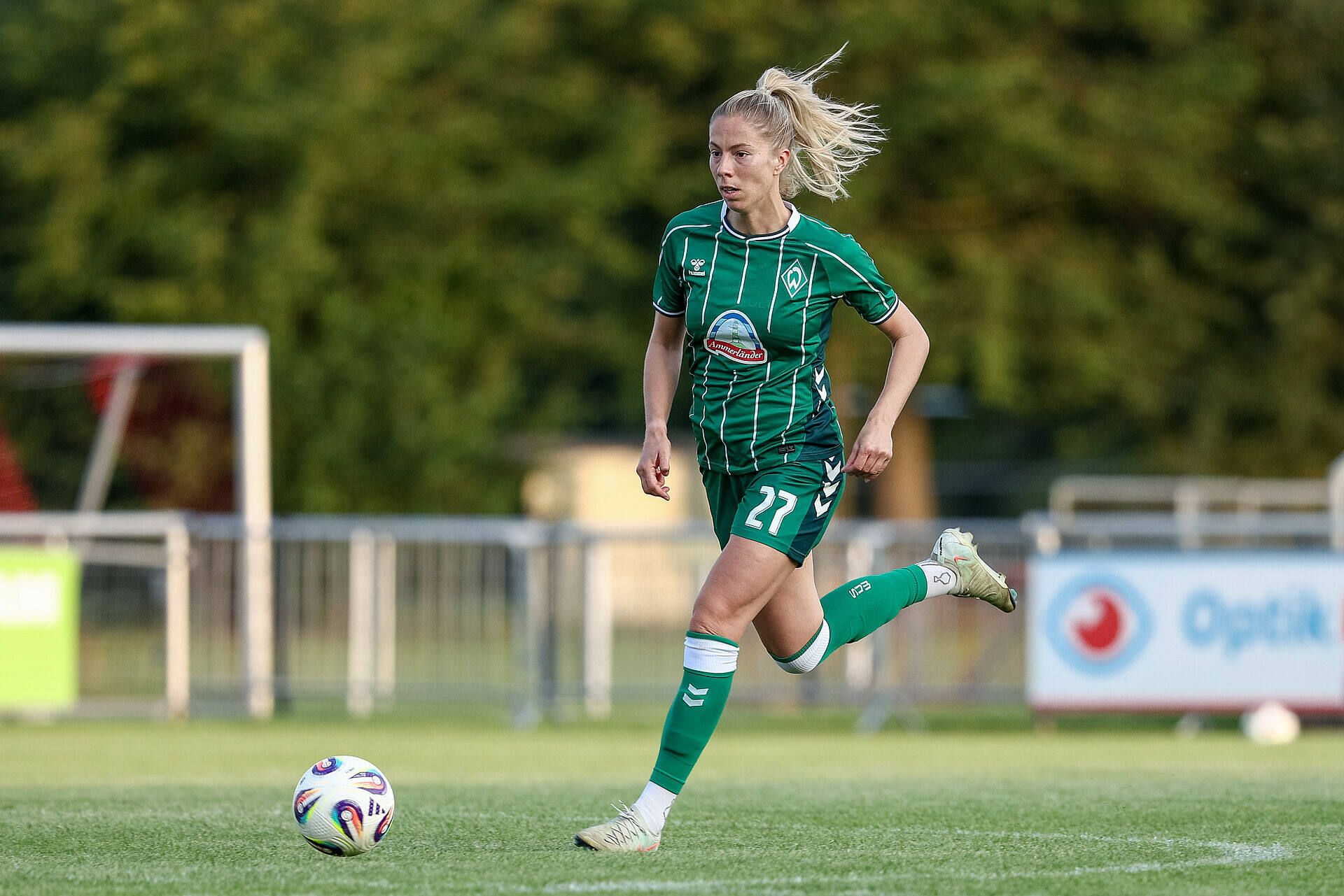 Sharon Beck dribbelt im grünen Trikot mit dem Ball auf dem Spielfeld, im Hintergrund sind Bäume und Zäune zu sehen.