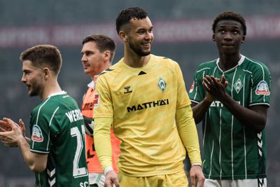 Ein Fußballspieler in einem gelben Trikot und ein Spieler in einem grünen Trikot, die auf dem Spielfeld lächeln und klatschen, vor dem Hintergrund eines verschwommenen Stadions.