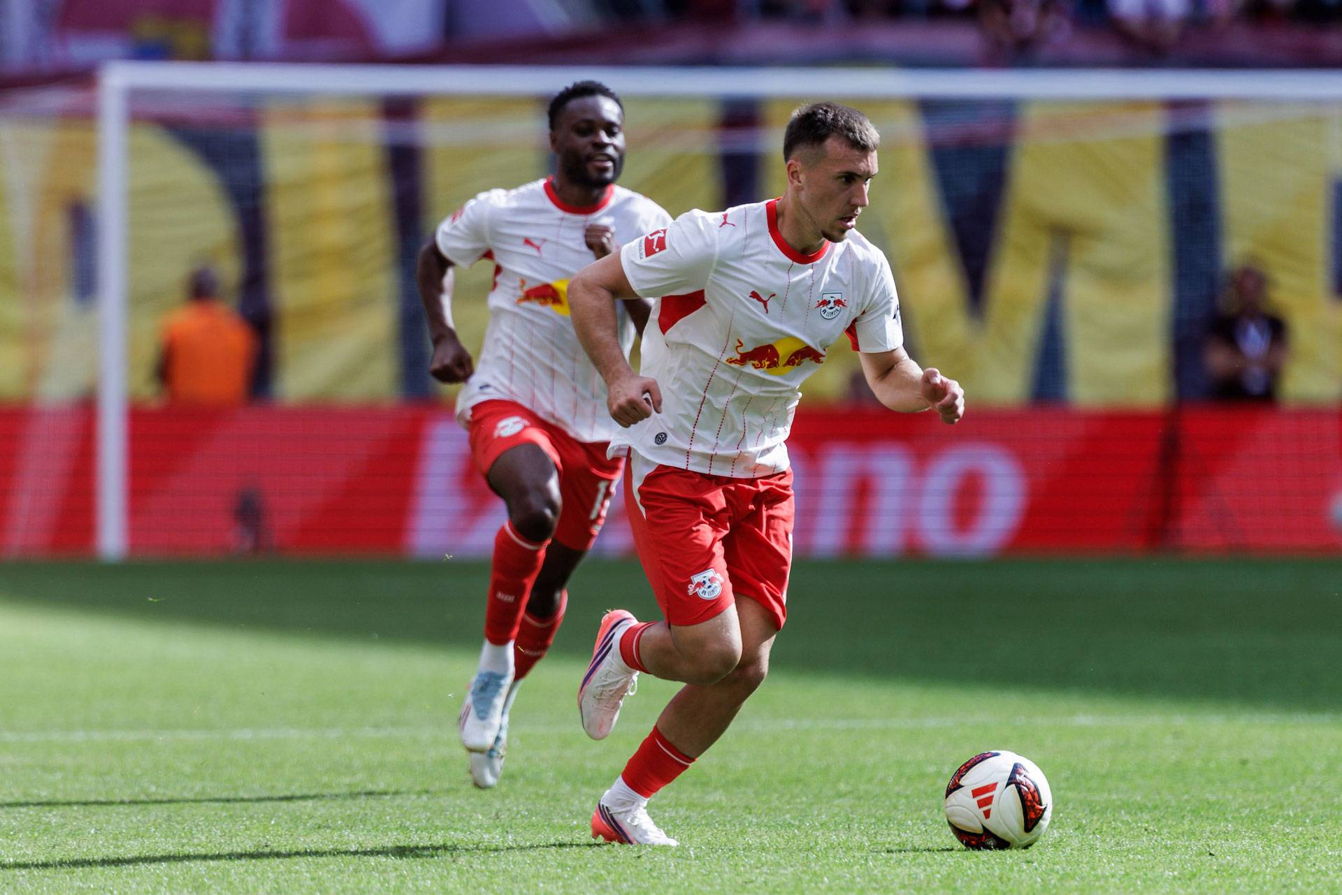 Zwei Fußballspieler in weißen und roten Trikots laufen über einen Rasenplatz, wobei sich ein Ball in der Nähe des Spielers im Vordergrund befindet.