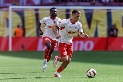 Zwei Fußballspieler in weißen und roten Trikots laufen über einen Rasenplatz, wobei sich ein Ball in der Nähe des Spielers im Vordergrund befindet.