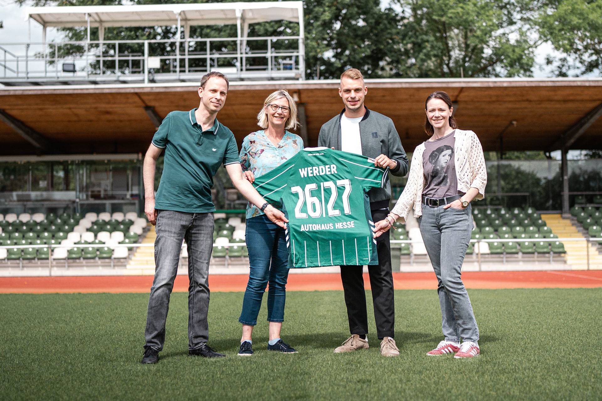 Vier Personen stehen auf einem Fußballfeld und halten ein grünes Werder-Trikot mit der Nummer 26127 in den Händen. Im Hintergrund sind leere Tribünen und Bäume zu sehen.
