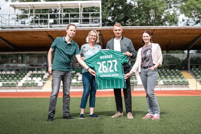 Vier Personen stehen auf einem Fußballfeld und halten ein grünes Werder-Trikot mit der Nummer 26127 in den Händen. Im Hintergrund sind leere Tribünen und Bäume zu sehen.