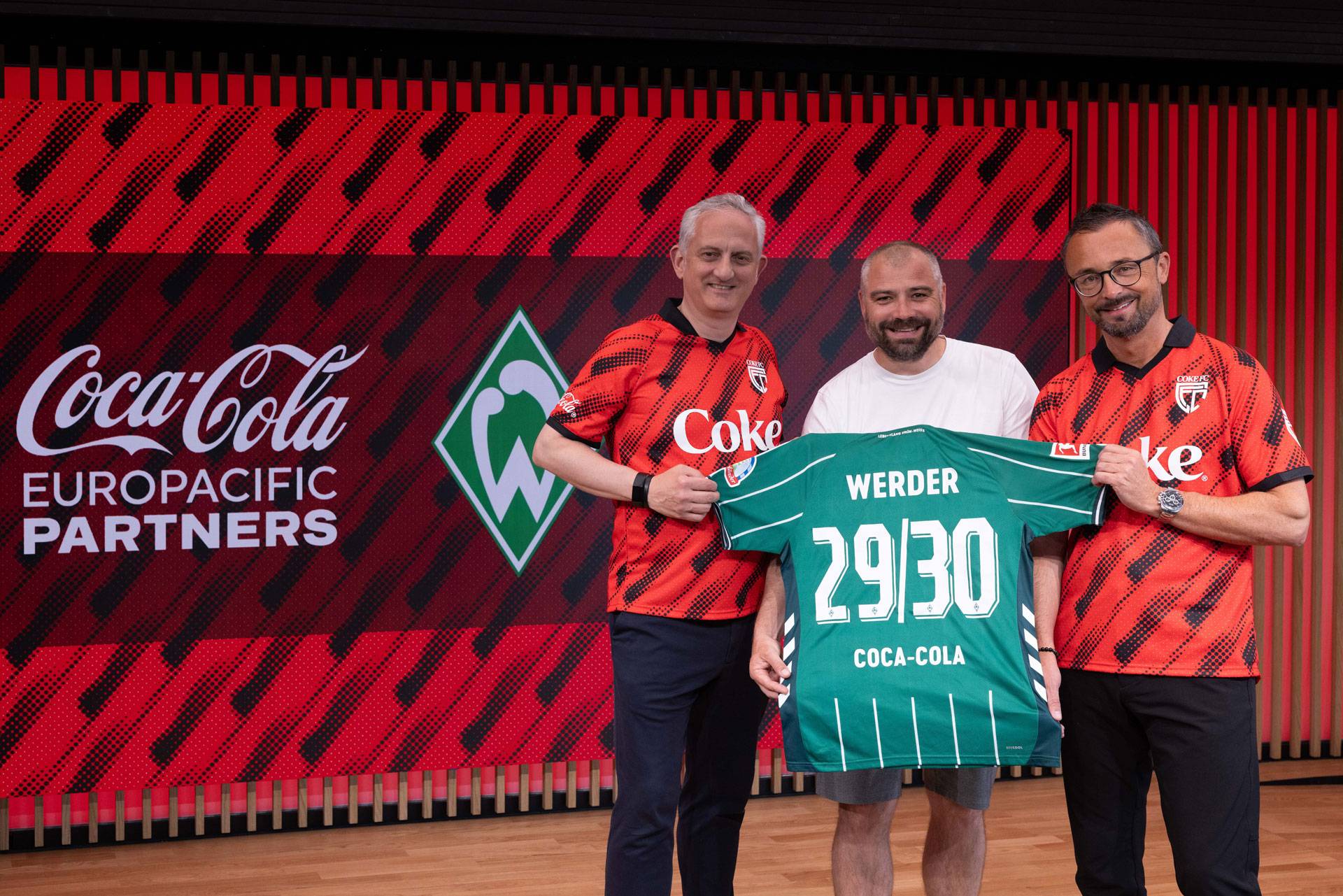 Drei Männer halten ein grünes Trikot von Werder Bremen mit der Aufschrift „29/30“ vor einem roten Hintergrund mit dem Logo von Coca-Cola Europacific Partners.