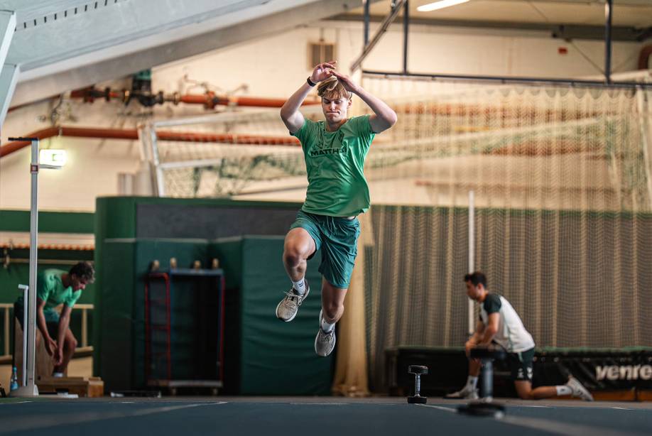 Ein Sportler in grüner Kleidung springt während des Hallentrainings, im Hintergrund sind Fitnessgeräte und ein Netz zu sehen. Andere beobachten ihn oder bereiten sich in der Nähe vor.