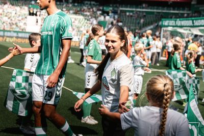 Fußballspieler und Kinder in grün-weißen Trikots begrüßen sich auf einem Fußballplatz, umgeben von Fahnen und Zuschauern.