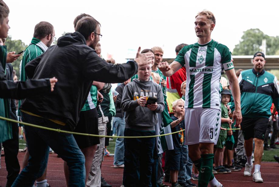 Ein Fußballspieler in einem grün-weißen Trikot winkt jubelnden Fans, darunter auch Kindern, entlang einer Absperrung bei einer Sportveranstaltung zu.