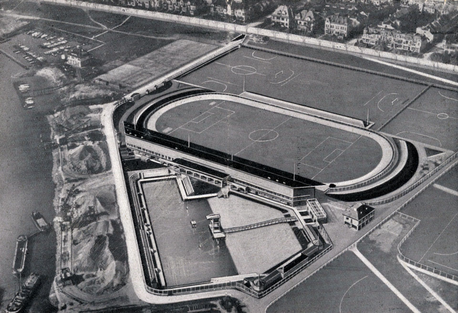 Schwarz-weiß Luftaufnahme eines ovalen Sportstadions mit angrenzenden Sportplätzen, umgeben von einem Wasserlauf und einem Wohngebiet.