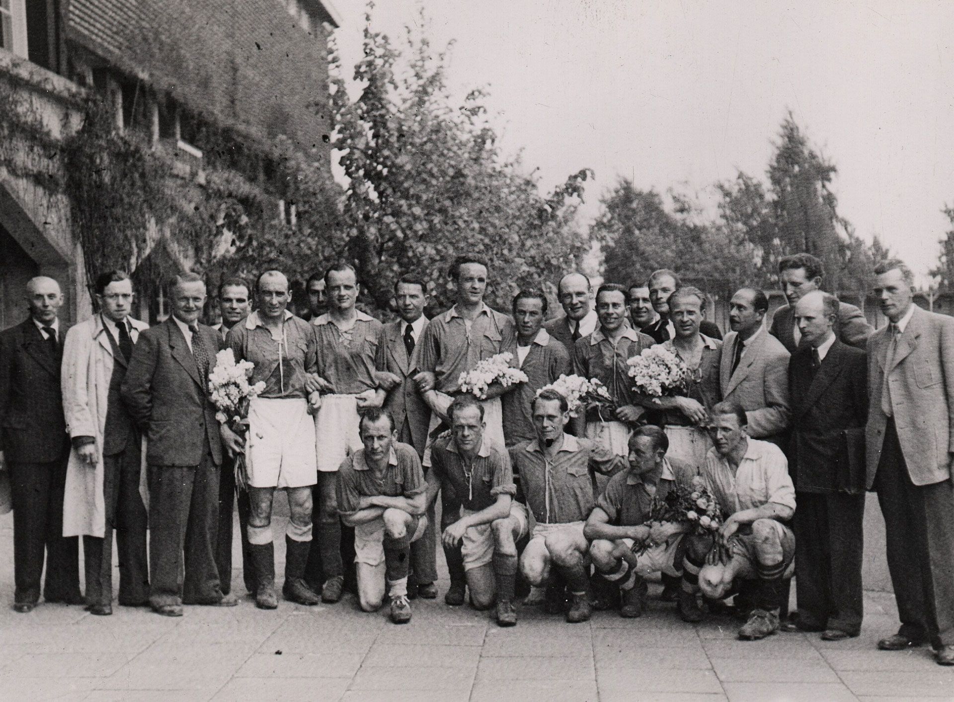 Ein Schwarz-Weiß-Foto einer alten Fußballmannschaft, die mit Blumensträußen posiert, umgeben von Männern in formeller Kleidung, vor dem Hintergrund von Bäumen und einem Gebäude.