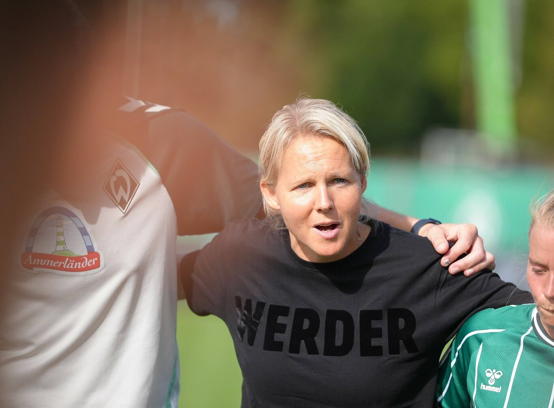 Trainerin Fritzy Kromp steht in einem schwarzen Pullover mit "WERDER"-Aufdruck im Mannschaftskreis und spricht.
