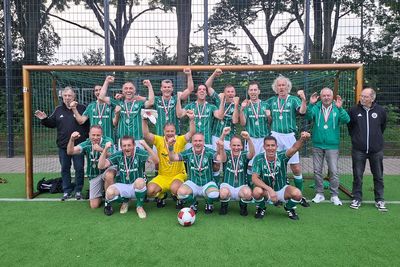 Eine Fußballmannschaft in grünen Trikots posiert mit Medaillen und feiert einen Sieg auf einem Spielfeld, im Hintergrund ist ein Tor mit Netz zu sehen.