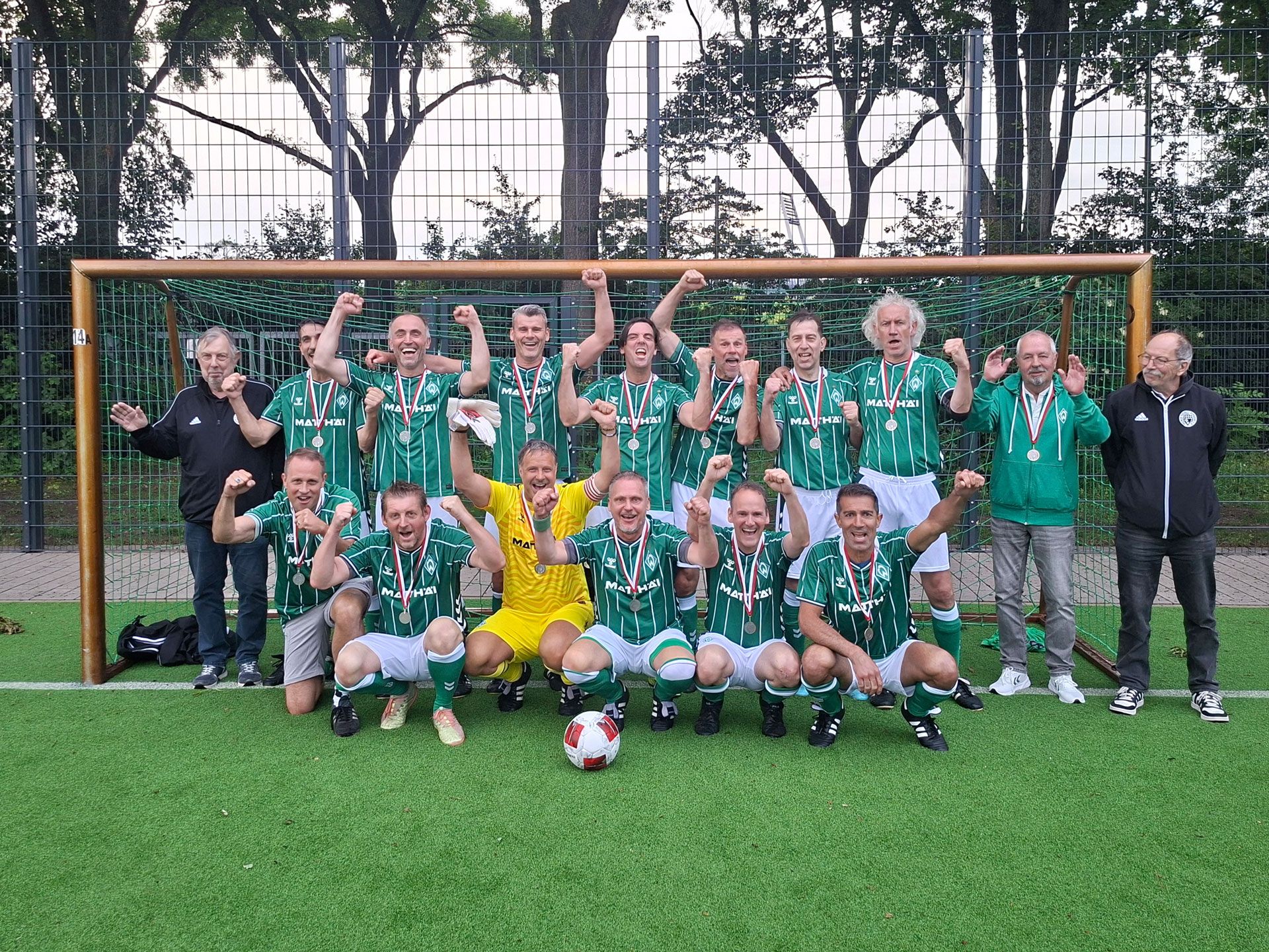 Eine Fußballmannschaft in grünen Trikots posiert mit Medaillen und feiert einen Sieg auf einem Spielfeld, im Hintergrund ist ein Tor mit Netz zu sehen.